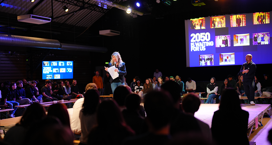 Woman speaking on stage with a microphone and holding papers during an indoor event, with an audience seated around her and a large screen in the background displaying '2050 is waiting for us