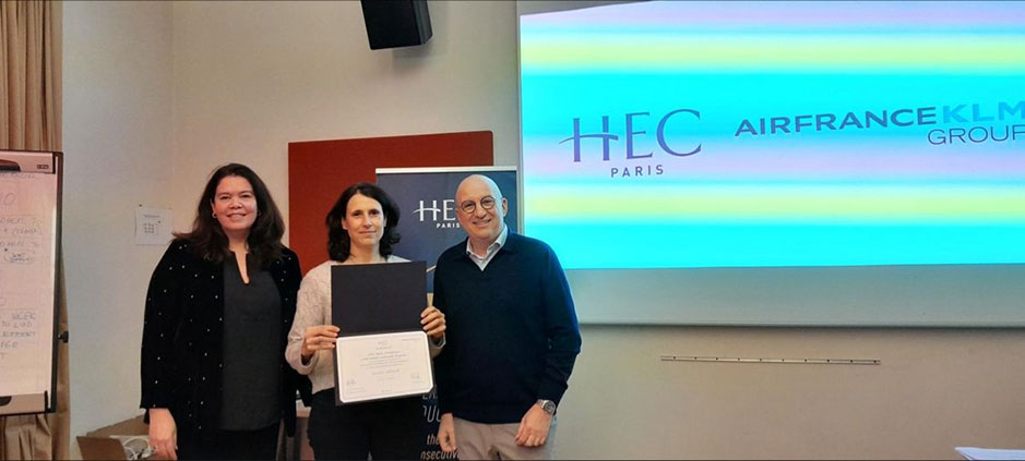 Three people stand in front of a screen displaying the HEC Paris and Air France KLM Group logos, with the woman in the center holding a certificate.