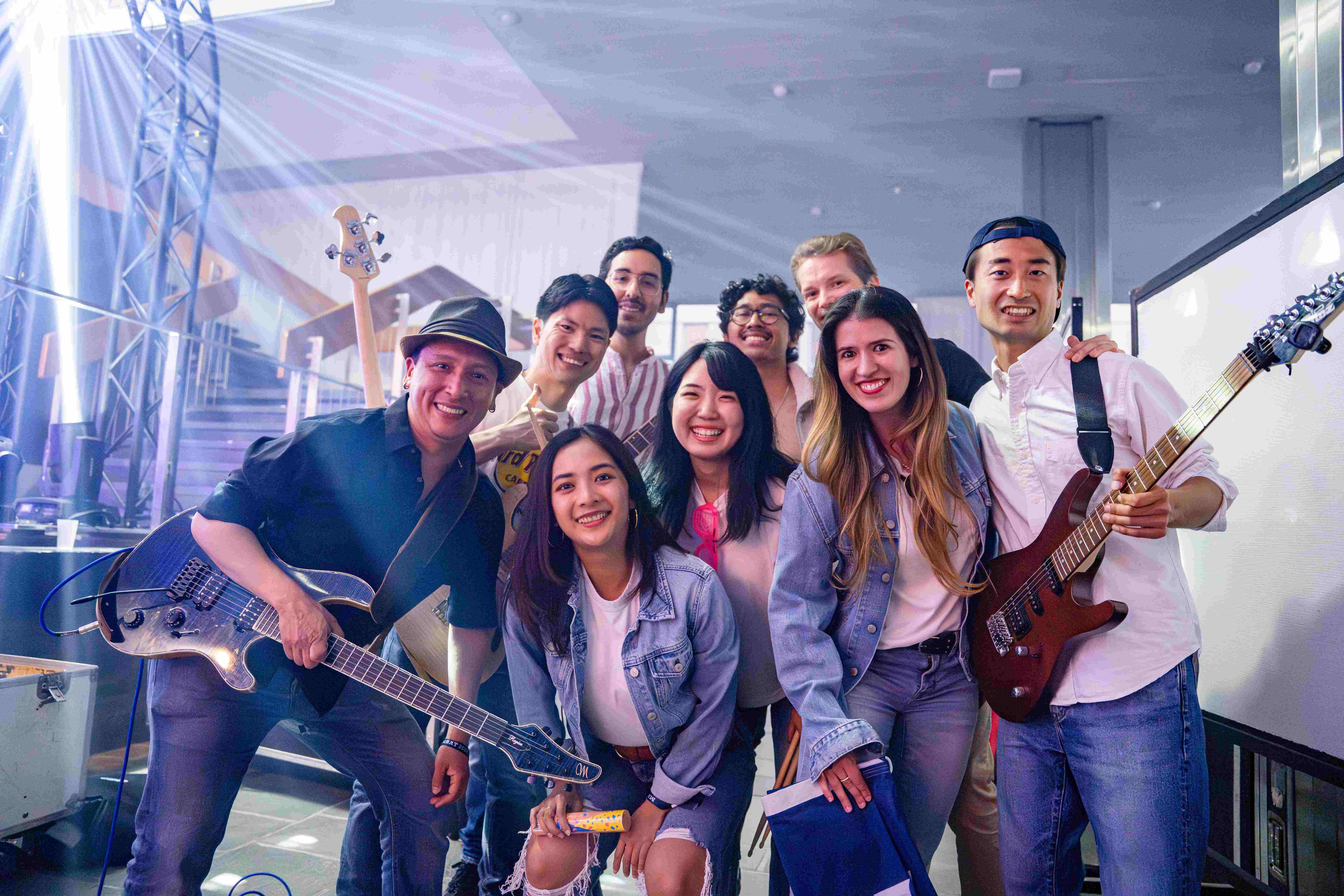 A group of nine cheerful people posing together, some holding musical instruments like guitars. They are in a lively indoor setting with stage lighting and equipment visible in the background, suggesting a musical performance or event