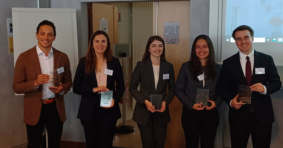 A group of five people standing indoors, each holding a plaque or award. They are dressed in formal business attire and are smiling for the camera. The background features a partially open door and a projection screen, suggesting a professional or academic setting, possibly an award ceremony or recognition event at HEC.