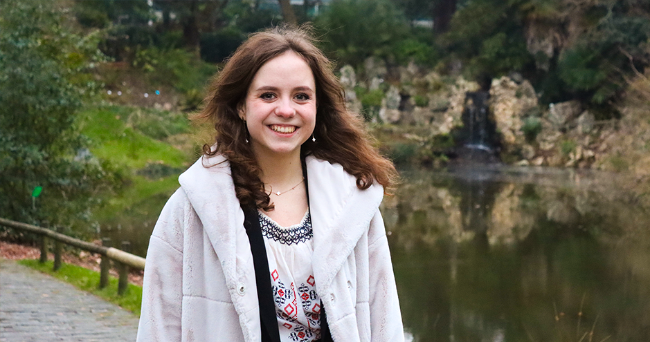 Une jeune femme souriante se tient sur un chemin pavé près d'un étang avec une petite cascade en arrière-plan. Elle porte un manteau blanc et un haut à motifs. Le cadre est naturel, avec des arbres et de la verdure autour