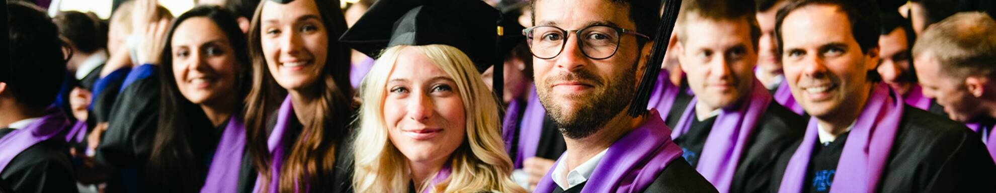 Un groupe de diplômés assis dans leurs toges et chapeaux académiques, souriant à la caméra. Ils portent des toges noires avec des écharpes violettes