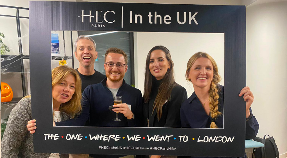 Five smiling people posing with a photo frame that reads HEC Paris In the UK - The One Where We Went to London' at an event in London