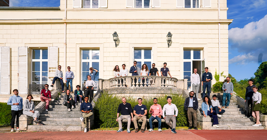 A group photo of around 30 people attending a workshop at HEC, posing on the steps of a large, elegant building with white walls and multiple windows